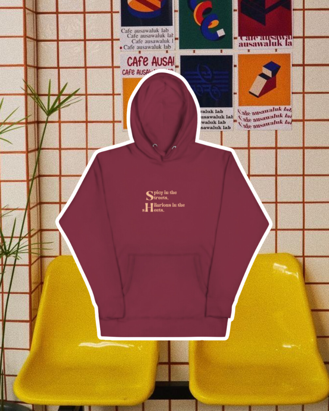 Maroon hoodie with the phrase 'Spicy in the streets, Hilarious in the sheets' on the chest, displayed against a vibrant café-style backdrop with yellow chairs and colorful posters. A playful and bold design for fans of funny merchandise and sarcasm.