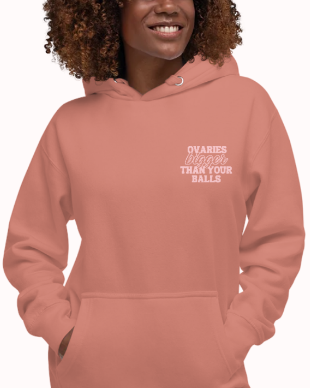 Front view of a dusty rose hoodie worn by a smiling woman, featuring the phrase 'Ovaries bigger than your balls' on the chest and back. Perfect for fans of funny merchandise, bold humor, and witty sarcasm.