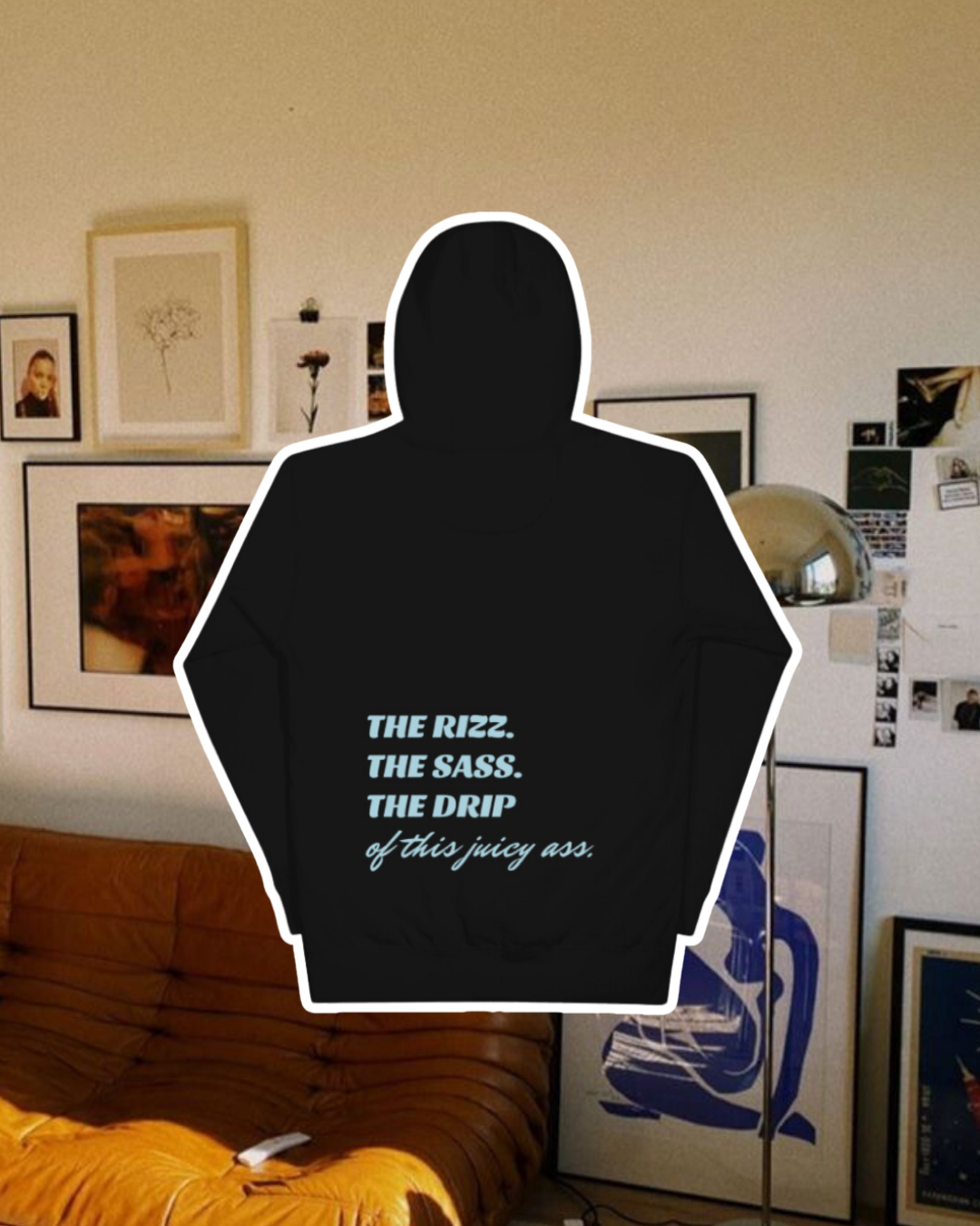 Black hoodie with the phrase 'The Rizz. The Sass. The Drip of this Juicy Ass' on the back, displayed against a vibrant living room with cozy elements and colorful frames. A playful and bold design for fans of funny merchandise and sarcasm.