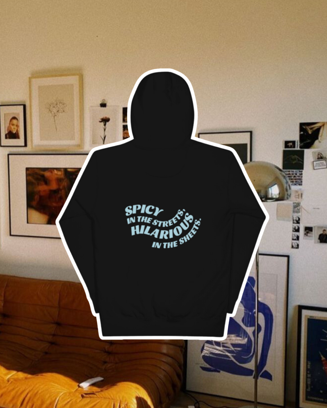 Black hoodie with the phrase 'Spicy in the streets, Hilarious in the sheets' on the back, displayed against a vibrant living room with cozy elements and colorful frames. A playful and bold design for fans of funny merchandise and sarcasm.