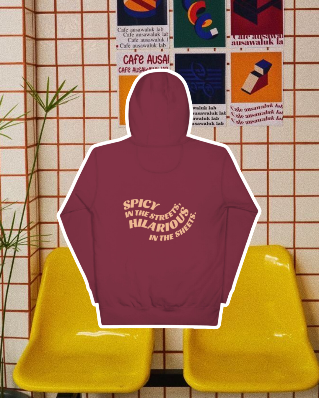 Maroon hoodie with the phrase 'Spicy in the streets, Hilarious in the sheets' on the back, displayed against a vibrant café-style backdrop with yellow chairs and colorful posters. A playful and bold design for fans of funny merchandise and sarcasm.