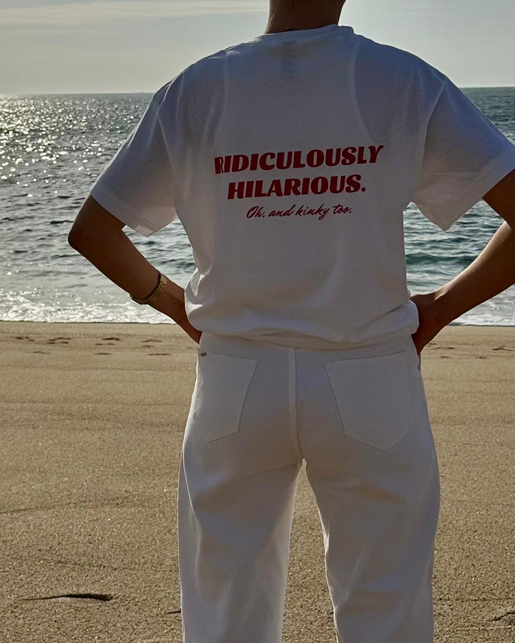 Woman using a white t-shirt with the phrase 'Ridiculously hilarious. Oh, and kinky too' on the back, at the beach. Perfect for fans of funny merchandise, bold humor, and witty sarcasm.