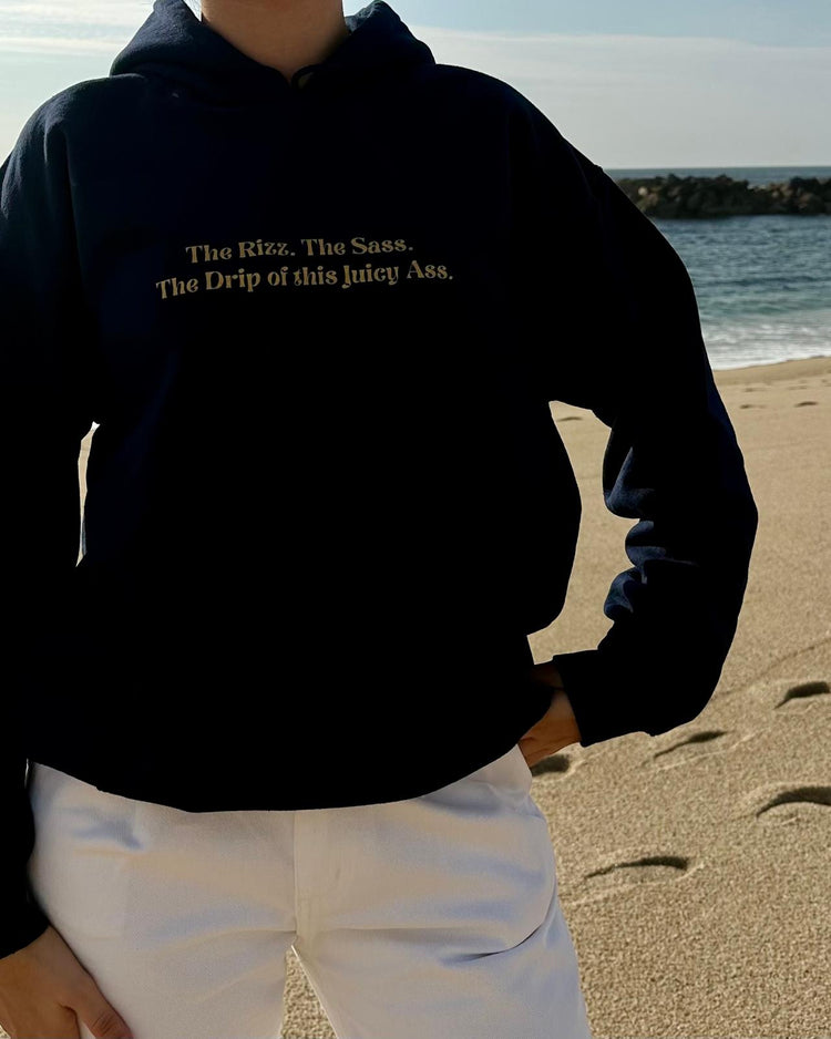 Woman using a navy hoodie with the phrase 'The Rizz. The Sass. The Drip of this Juicy Ass' on the chest, at the beach. Perfect for fans of funny merchandise, bold humor, and witty sarcasm.