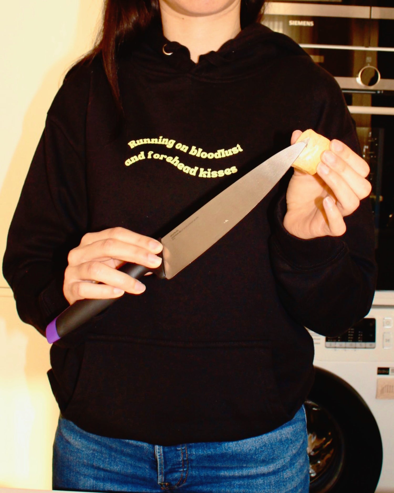 Woman in a black hoodie that reads "Running on bloodlust and forehead kisses" while using an 8-inch chef's knife to spread butter on a toast. A blend of domesticity and dark humor, perfect for fans of ironic fashion and bold statement apparel.
