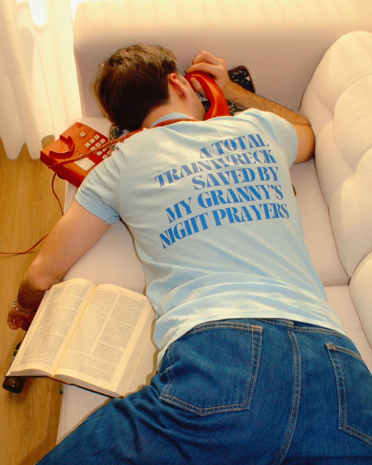 Man lying on a couch, wearing a light blue t-shirt that reads "A total trainwreck saved by my granny’s night prayers." A red rotary phone and an open book are beside him. Perfect scene for fans of funny merchandise, bold humor, and witty sarcasm.
