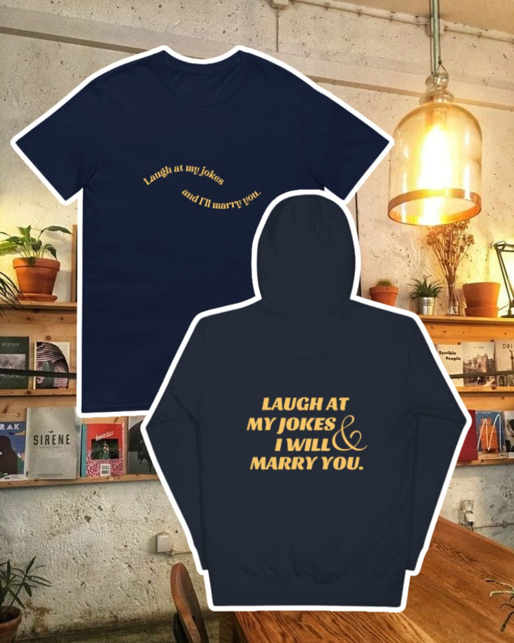 Navy blazer hoodie with the phrase 'Laugh at my jokes and I will marry you' on the back, displayed against a vibrant café-style backdrop with wood chairs and colorful cozy decoration. A playful and bold design for fans of funny merchandise and sarcasm.