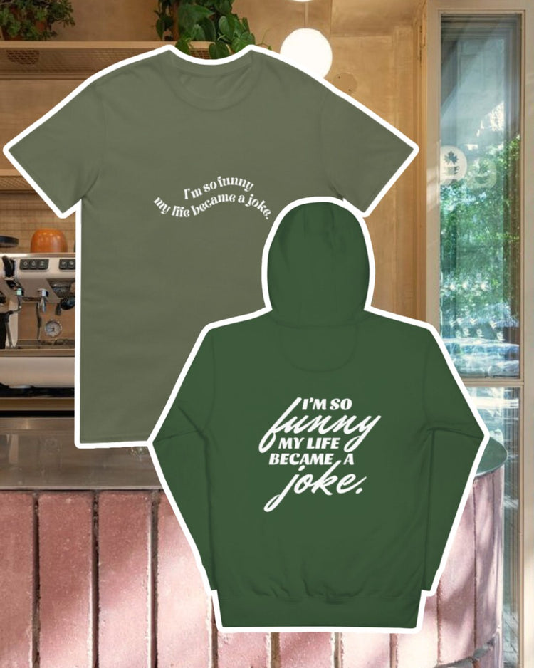 Forest green hoodie with the phrase 'I'm so funny my life became a joke' on the back, displayed against a vibrant café-style backdrop with a pink counter and cozy decoration with plants. A playful and bold design for fans of funny merchandise and sarcasm.