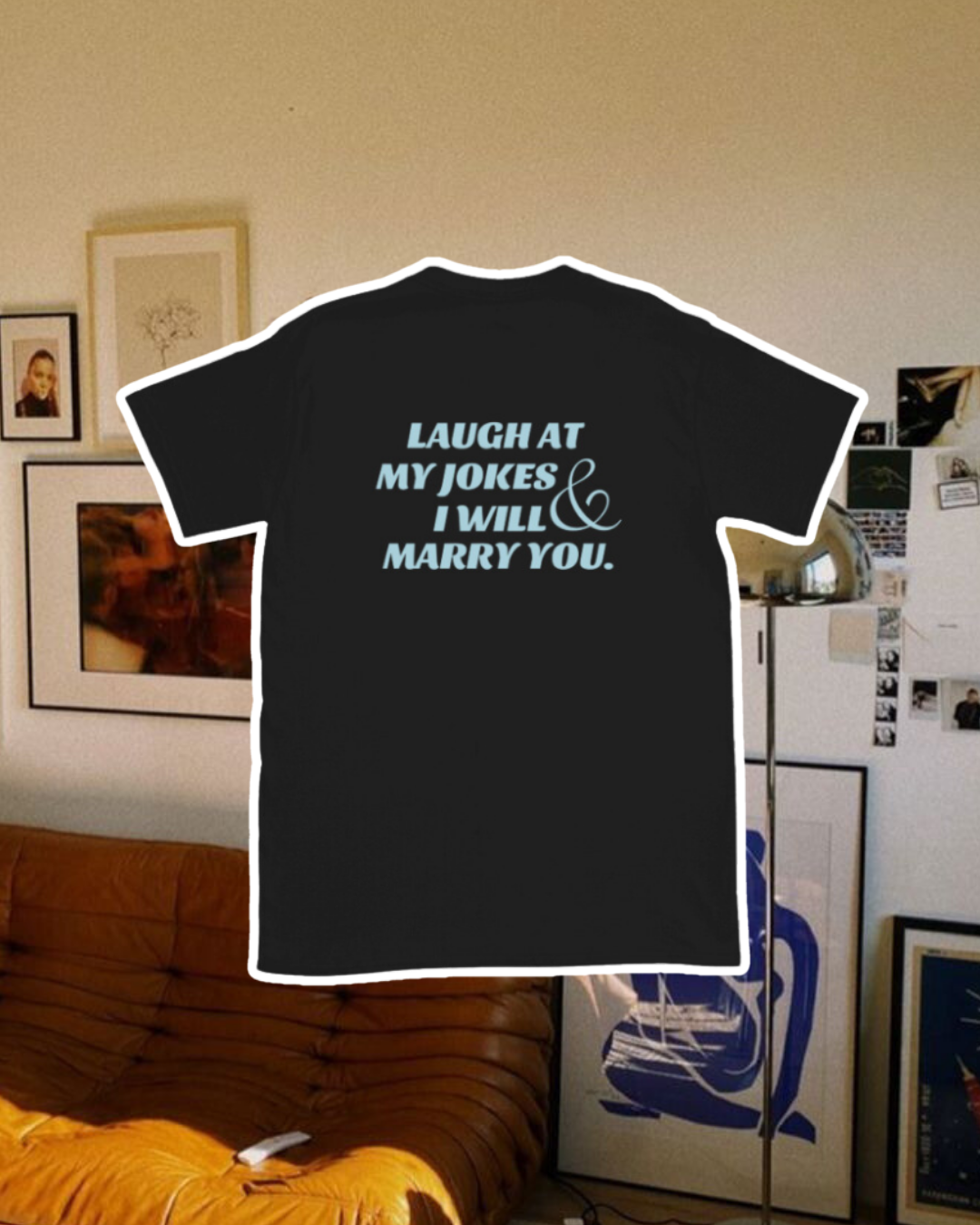 Black T-Shirt with the phrase 'Laugh at my jokes and I will marry you' on the back, displayed against a vibrant living room with cozy elements and colorful frames. A playful and bold design for fans of funny merchandise and sarcasm.