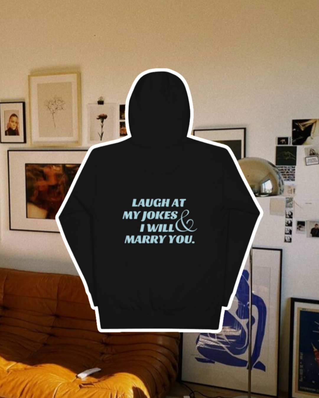 Black hoodie with the phrase 'Laugh at my jokes and I will marry you' on the back, displayed against a vibrant living room with cozy elements and colorful frames. A playful and bold design for fans of funny merchandise and sarcasm.