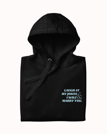 Folded black hoodie featuring the ironic phrase 'Laugh at my jokes and I will marry you'. Perfect for fans of funny merchandise, bold humor, and witty sarcasm.