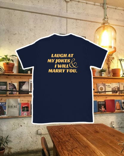 Navy T-Shirt with the phrase 'Laugh at my jokes and I will marry you' on the back, displayed against a vibrant café-style backdrop with wood chairs and colorful cozy decoration. A playful and bold design for fans of funny merchandise and sarcasm.