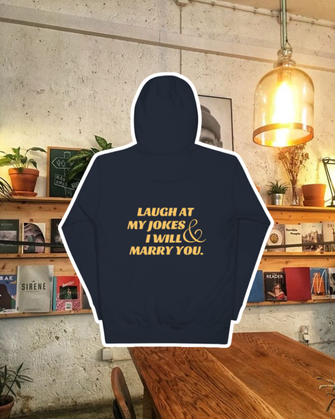 Navy blazer hoodie with the phrase 'Laugh at my jokes and I will marry you' on the back, displayed against a vibrant café-style backdrop with wood chairs and colorful cozy decoration. A playful and bold design for fans of funny merchandise and sarcasm.