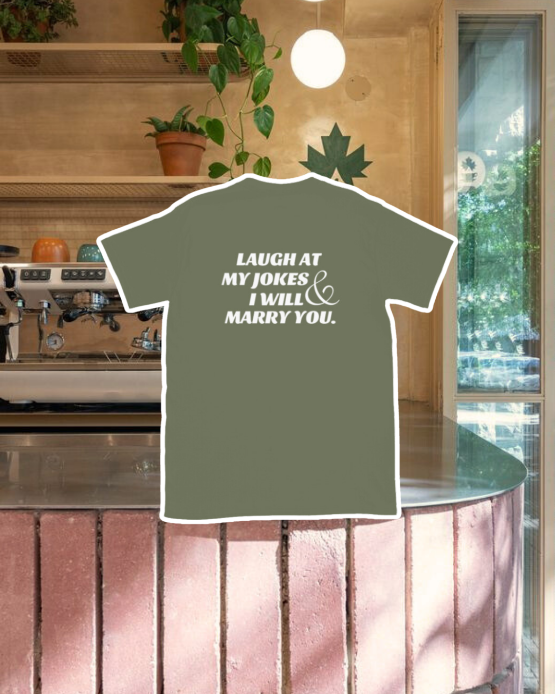 Military green T-Shirt with the phrase 'Laugh at my jokes and I will marry you' on the back, displayed against a vibrant café-style backdrop with a pink counter and cozy decoration with plants. A playful and bold design for fans of funny merchandise and sarcasm.