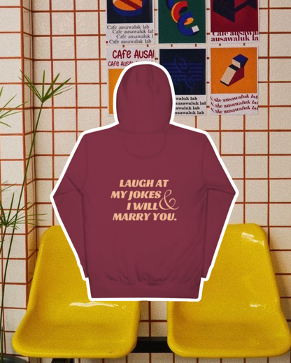 Maroon hoodie with the phrase 'Laugh at my jokes and I will marry you' on the back, displayed against a vibrant café-style backdrop with yellow chairs and colorful posters. A playful and bold design for fans of funny merchandise and sarcasm. 