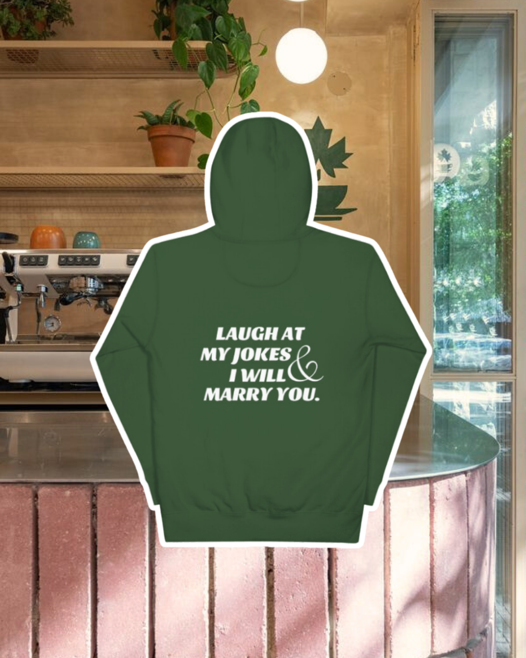 Forest green hoodie with the phrase 'Laugh at my jokes and I will marry you' on the back, displayed against a vibrant café-style backdrop with a pink counter and cozy decoration with plants. A playful and bold design for fans of funny merchandise and sarcasm.