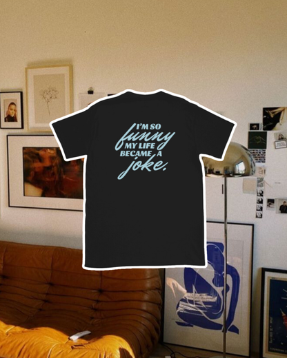 Black T-Shirt with the phrase 'I'm so funny my life became a joke' on the back, displayed against a vibrant living room with cozy elements and colorful frames. A playful and bold design for fans of funny merchandise and sarcasm.