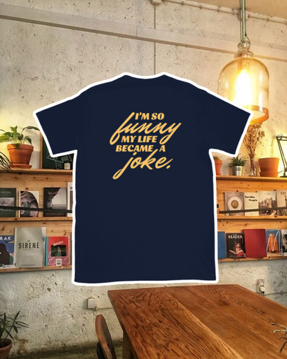 Navy T-Shirt with the phrase 'I'm so funny my life became a joke' on the back, displayed against a vibrant café-style backdrop with wood chairs and colorful cozy decoration. A playful and bold design for fans of funny merchandise and sarcasm.