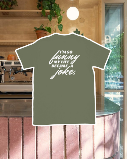 Military green T-Shirt with the phrase 'I'm so funny my life became a joke' on the back, displayed against a vibrant café-style backdrop with a pink counter and cozy decoration with plants. A playful and bold design for fans of funny merchandise and sarcasm.