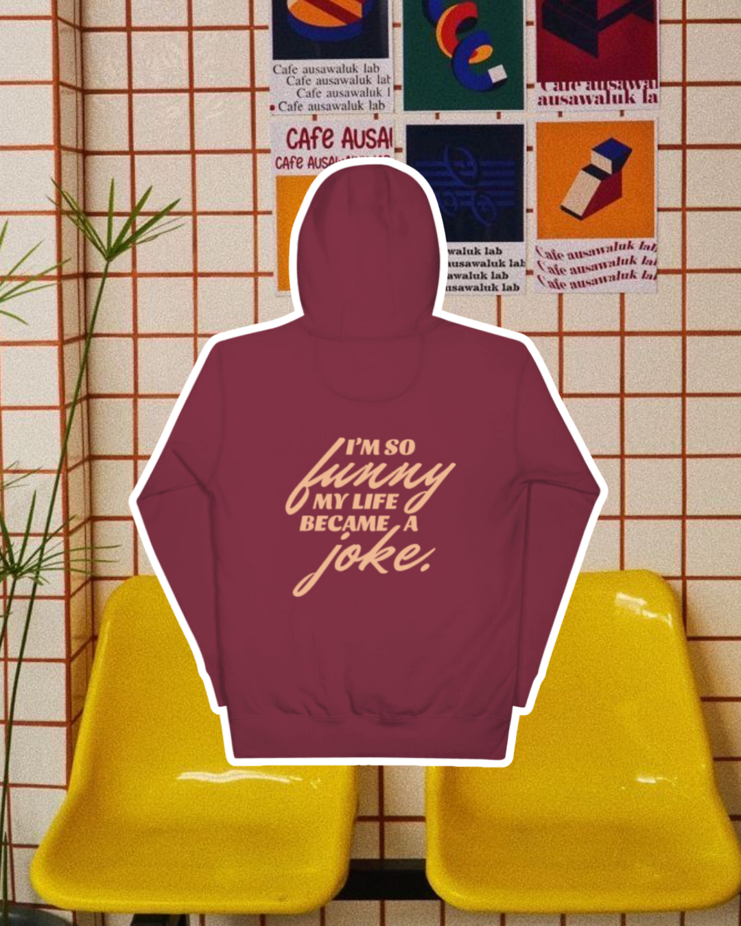 Maroon hoodie with the phrase 'I'm so funny my life became a joke' on the back, displayed against a vibrant café-style backdrop with yellow chairs and colorful posters. A playful and bold design for fans of funny merchandise and sarcasm.