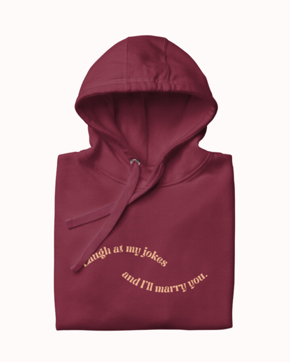 Folded maroon hoodie featuring the ironic phrase 'Laugh at my jokes and I'll marry you'. Perfect for fans of funny merchandise, bold humor, and witty sarcasm. 