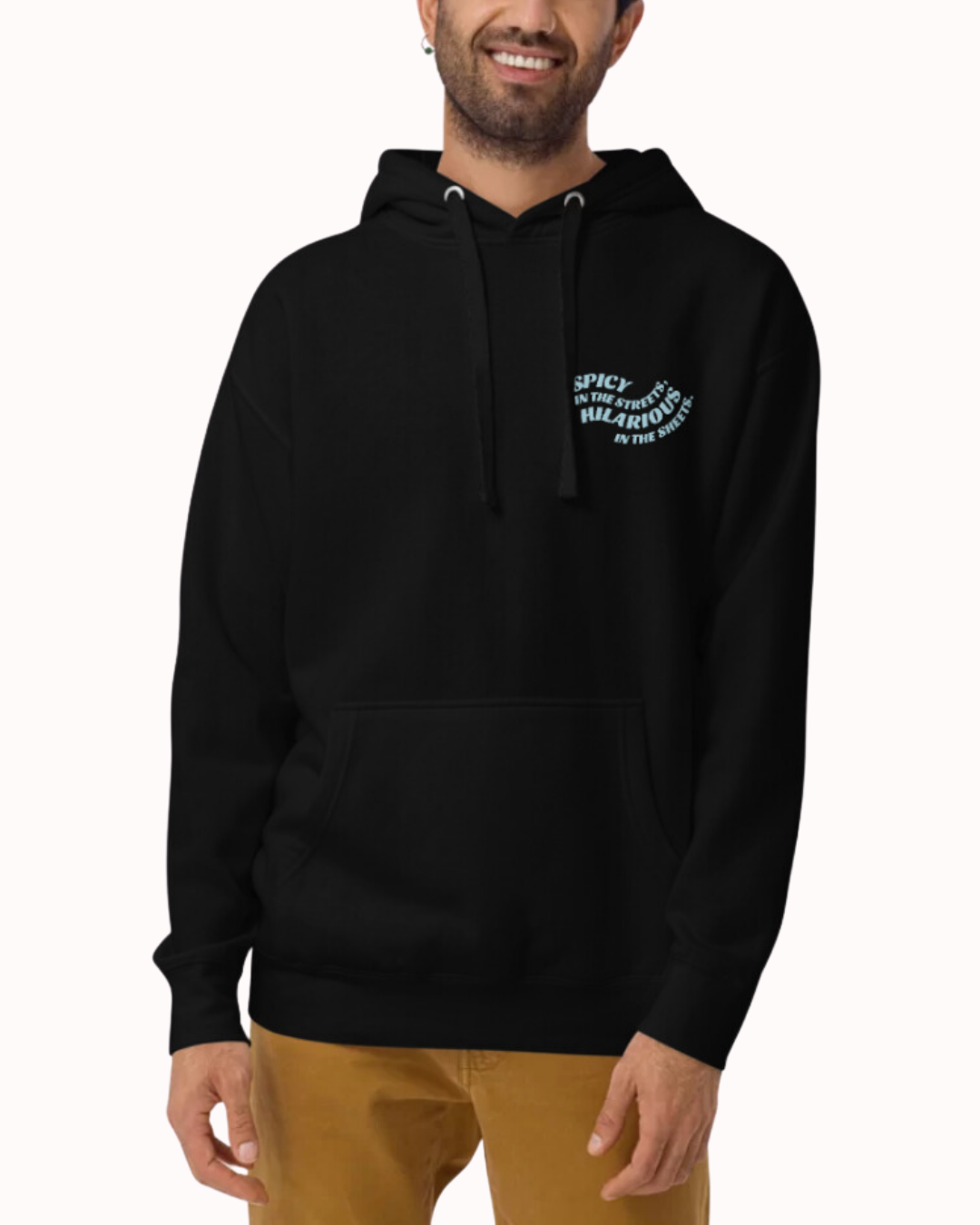 Front view of a black hoodie worn by a smiling man, featuring the phrase 'Spicy in the streets, Hilarious in the sheets' on the chest and back. Perfect for fans of funny merchandise, bold humor, and witty sarcasm.