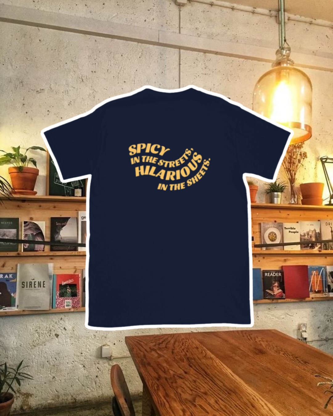 Navy blue T-Shirt with the phrase 'Spicy in the streets, Hilarious in the sheets' on the back, displayed against a vibrant café-style backdrop with wood chairs and colorful cozy decoration. A playful and bold design for fans of funny merchandise and sarcasm.