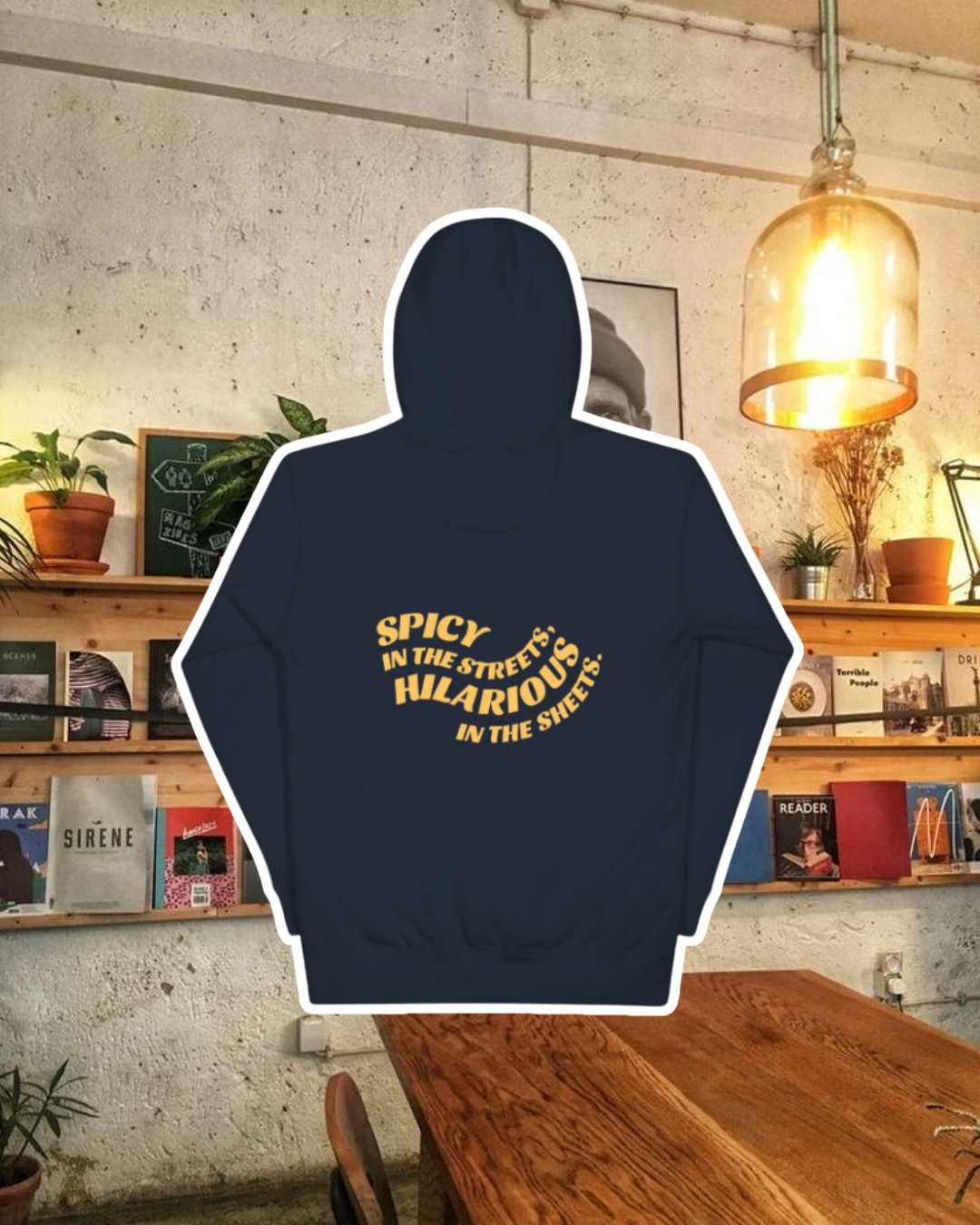 Navy blazer hoodie with the phrase 'Spicy in the streets, Hilarious in the sheets' on the back, displayed against a vibrant café-style backdrop with wood chairs and colorful cozy decoration. A playful and bold design for fans of funny merchandise and sarcasm.