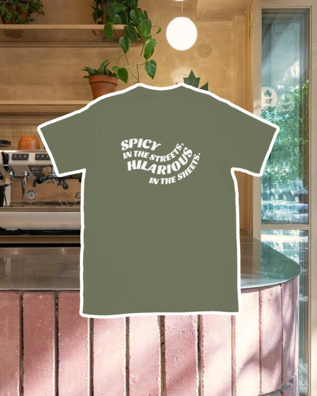 Military green T-Shirt with the phrase 'Spicy in the streets, Hilarious in the sheets' on the back, displayed against a vibrant café-style backdrop with a pink counter and cozy decoration with plants. A playful and bold design for fans of funny merchandise and sarcasm.