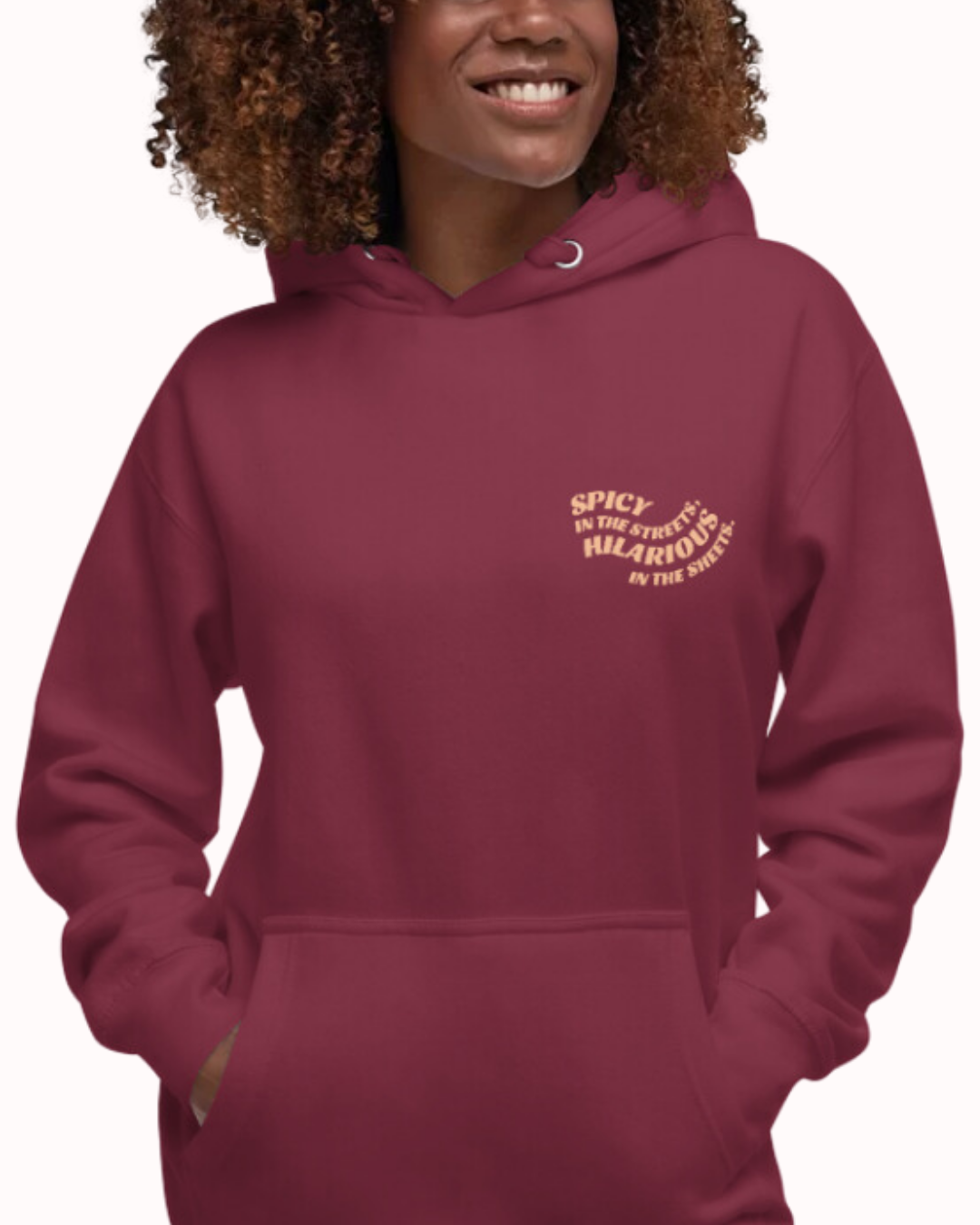 Front view of a maroon hoodie worn by a smiling woman, featuring the phrase 'Spicy in the streets, Hilarious in the sheets' on the chest and back. Perfect for fans of funny merchandise, bold humor, and witty sarcasm.