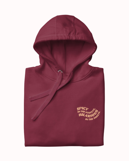 Folded maroon hoodie featuring the ironic phrase 'Spicy in the streets, Hilarious in the sheets'. Perfect for fans of funny merchandise, bold humor, and witty sarcasm. 