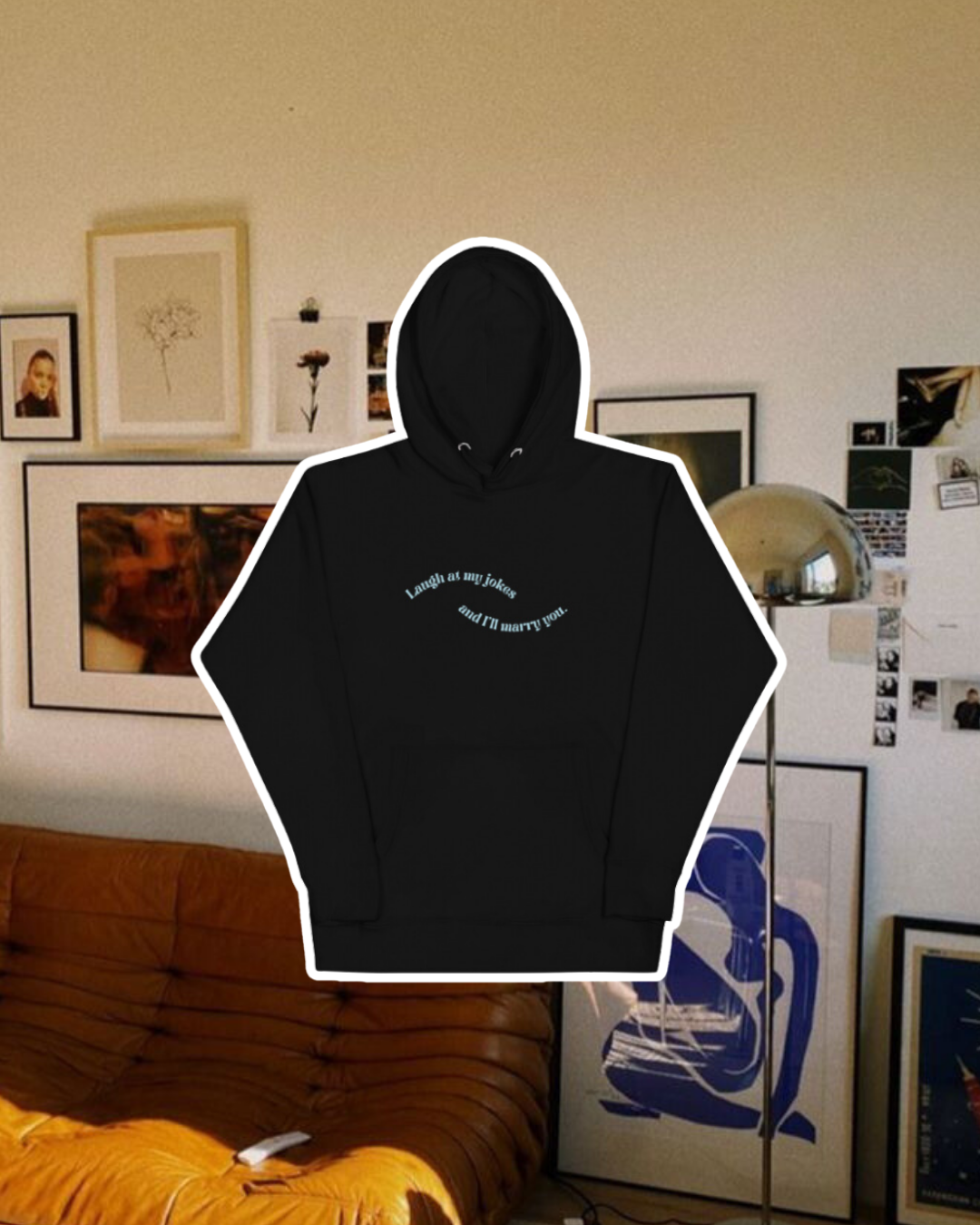 Black hoodie with the phrase 'Laugh at my jokes and I'll marry you' on the chest, displayed against a vibrant living room with cozy elements and colorful frames. A playful and bold design for fans of funny merchandise and sarcasm.