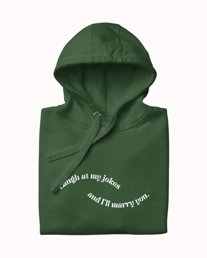 Folded forest green hoodie featuring the ironic phrase 'Laugh at my jokes and I'll marry you'. Perfect for fans of funny merchandise, bold humor, and witty sarcasm.