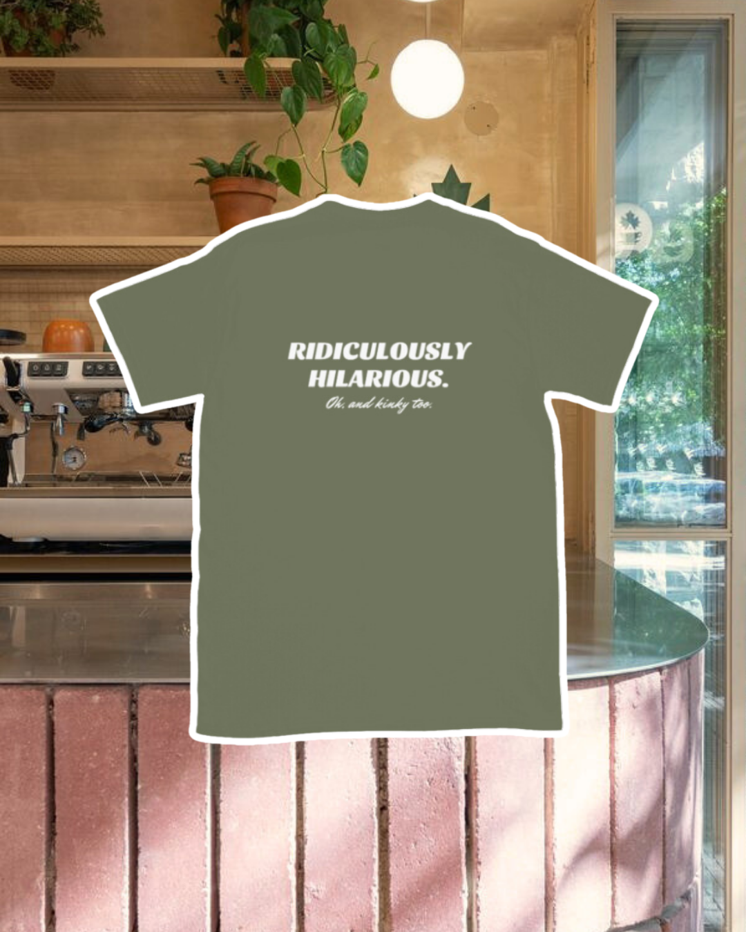 Military green T-Shirt with the phrase 'Ridiculously hilarious. Oh, and kinky too' on the back, displayed against a vibrant café-style backdrop with a pink counter and cozy decoration with plants. A playful and bold design for fans of funny merchandise and sarcasm.