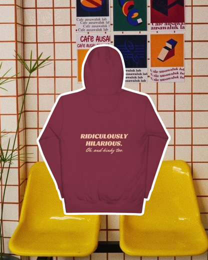 Maroon hoodie with the phrase 'Ridiculously hilarious. Oh, and kinky too' on the back, displayed against a vibrant café-style backdrop with yellow chairs and colorful posters. A playful and bold design for fans of funny merchandise and sarcasm.