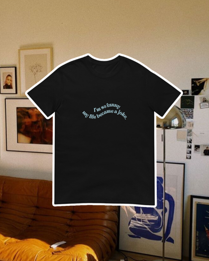 Black T-Shirt with the phrase 'I'm so funny my life became a joke' on the chest, displayed against a vibrant living room with cozy elements and colorful frames. A playful and bold design for fans of funny merchandise and sarcasm.