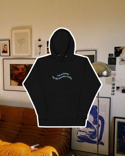 Black hoodie with the phrase 'I'm so funny my life became a joke' on the chest, displayed against a vibrant living room with cozy elements and colorful frames. A playful and bold design for fans of funny merchandise and sarcasm.