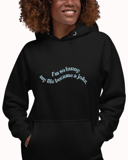 Front view of a black hoodie worn by a smiling woman, featuring the phrase 'I'm so funny my life became a joke' on the chest. Perfect for fans of funny merchandise, bold humor, and witty sarcasm.