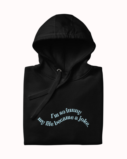 Folded black hoodie featuring the ironic phrase 'I'm so funny my life became a joke'. Perfect for fans of funny merchandise, bold humor, and witty sarcasm.