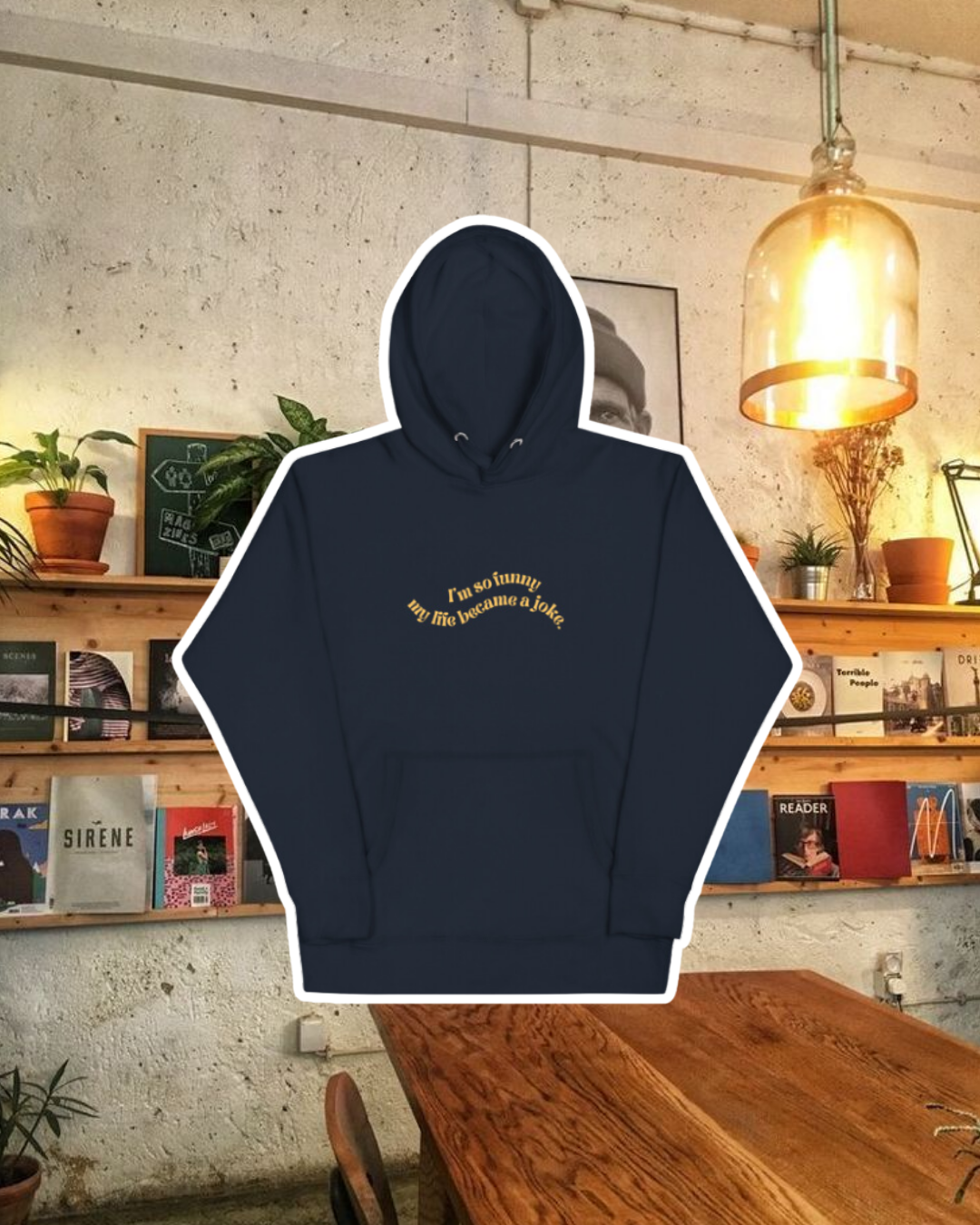 Navy blazer hoodie with the phrase 'I'm so funny my life became a joke' on the chest, displayed against a vibrant café-style backdrop with wood chairs and colorful cozy decoration. A playful and bold design for fans of funny merchandise and sarcasm.