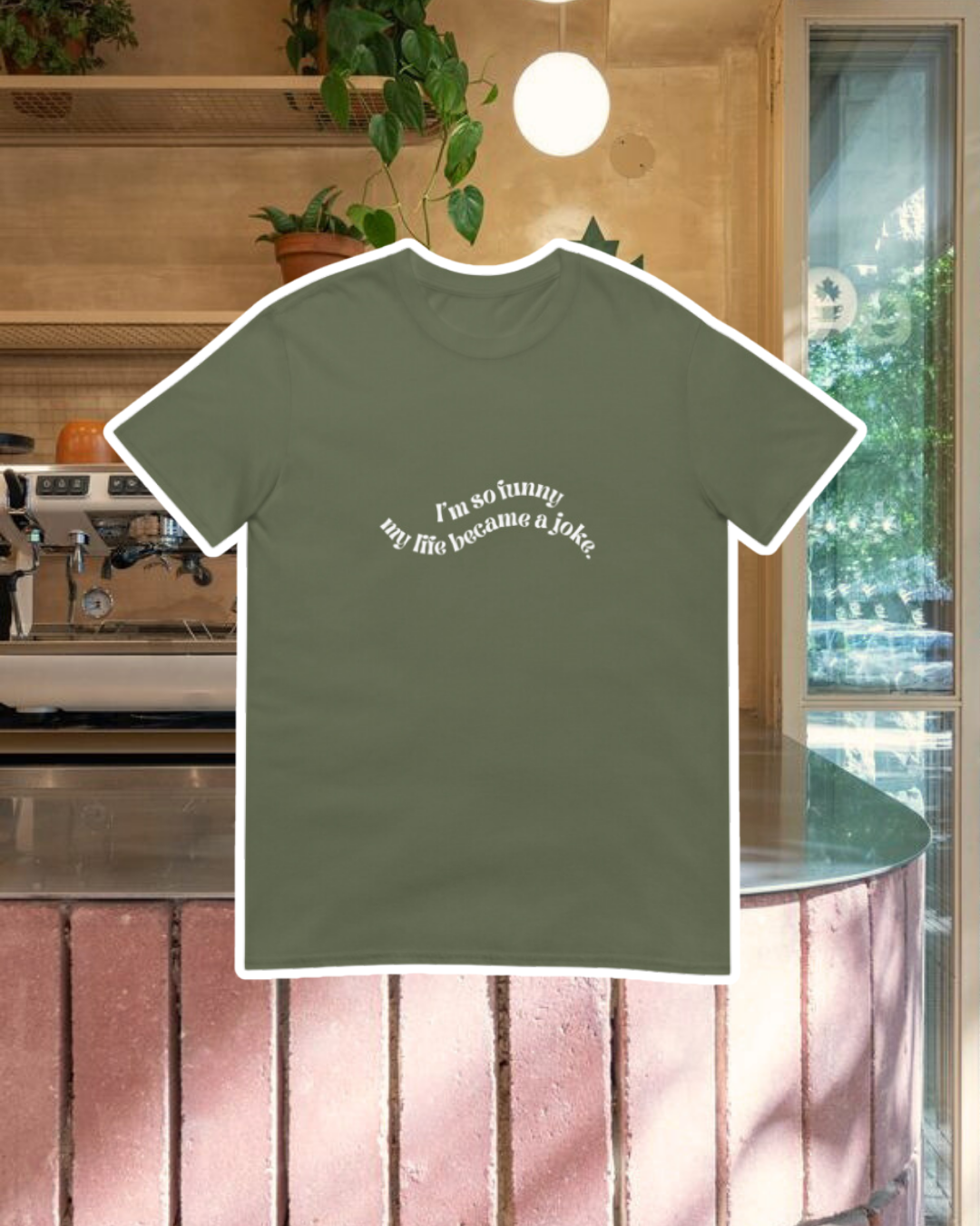 Military green T-Shirt with the phrase 'I'm so funny my life became a joke' on the chest, displayed against a vibrant café-style backdrop with wood chairs and colorful cozy decoration. A playful and bold design for fans of funny merchandise and sarcasm.