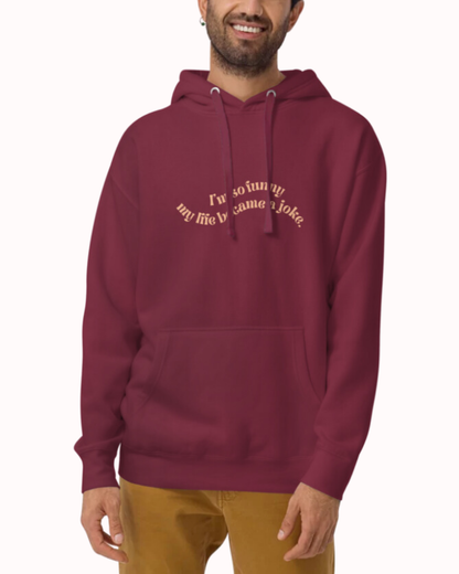 Front view of a maroon hoodie worn by a smiling man, featuring the phrase 'I'm so funny my life became a joke' on the chest. Perfect for fans of funny merchandise, bold humor, and witty sarcasm. 