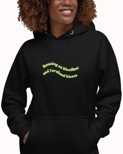 Front view of a Black hoodie worn by a smiling woman, featuring the phrase "Running on bloodlust and forehead kisses" on the chest and back. Perfect for fans of funny merchandise, bold humor, and witty sarcasm.