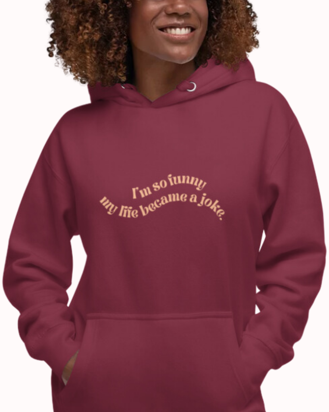 Front view of a maroon hoodie worn by a smiling woman, featuring the phrase 'I'm so funny my life became a joke' on the chest. Perfect for fans of funny merchandise, bold humor, and witty sarcasm.