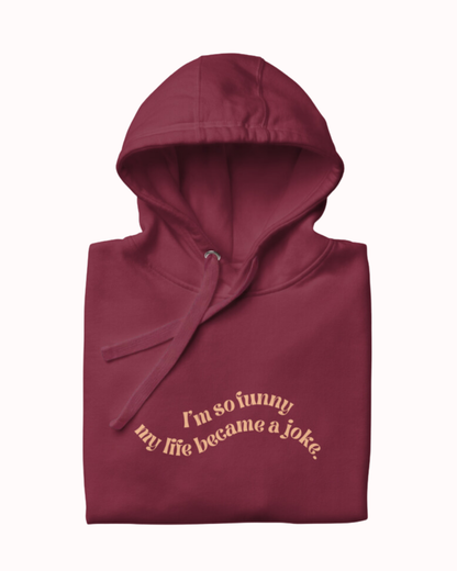 Folded maroon hoodie featuring the ironic phrase 'I'm so funny my life became a joke'. Perfect for fans of funny merchandise, bold humor, and witty sarcasm. 