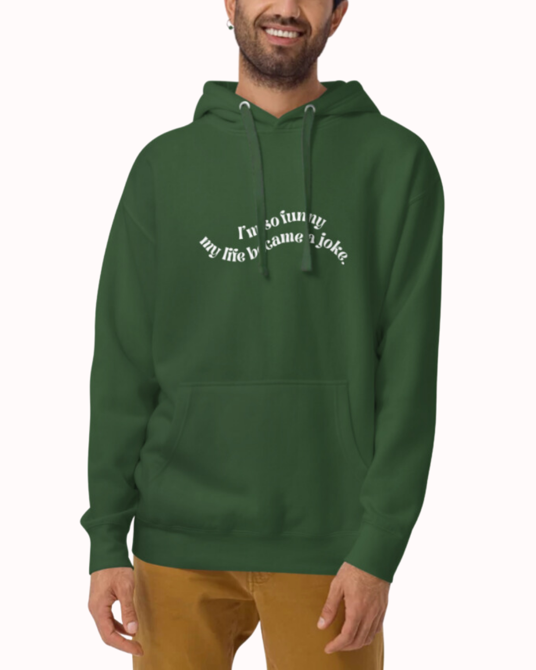 Front view of a forest green hoodie worn by a smiling man, featuring the phrase 'I'm so funny my life became a joke' on the chest. Perfect for fans of funny merchandise, bold humor, and witty sarcasm.