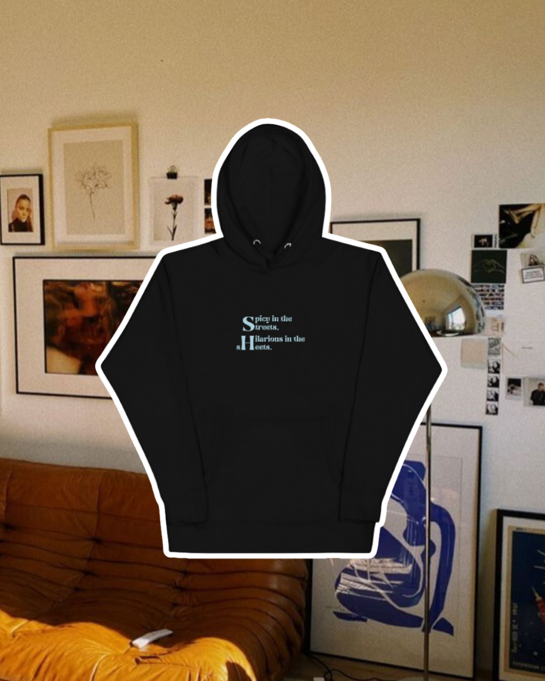 Black hoodie with the phrase 'Spicy in the streets, Hilarious in the sheets' on the chest, displayed against a vibrant living room with cozy elements and colorful frames. A playful and bold design for fans of funny merchandise and sarcasm.