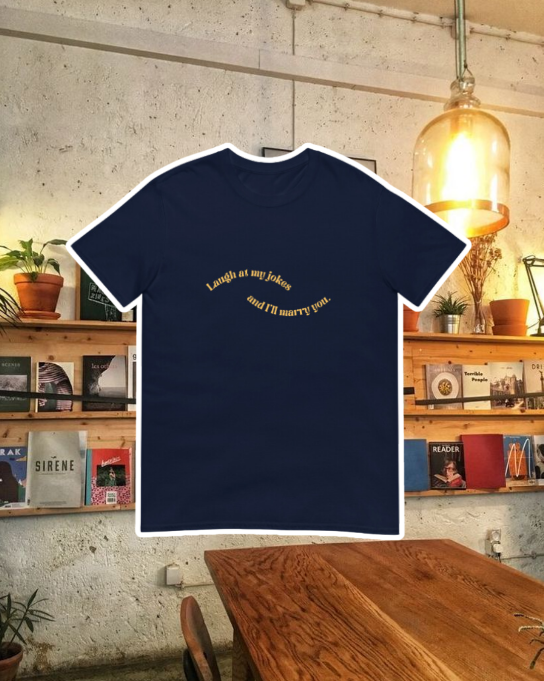 Navy T-Shirt with the phrase 'Laugh at my jokes and I will marry you' on the chest, displayed against a vibrant café-style backdrop with wood chairs and colorful cozy decoration. A playful and bold design for fans of funny merchandise and sarcasm.