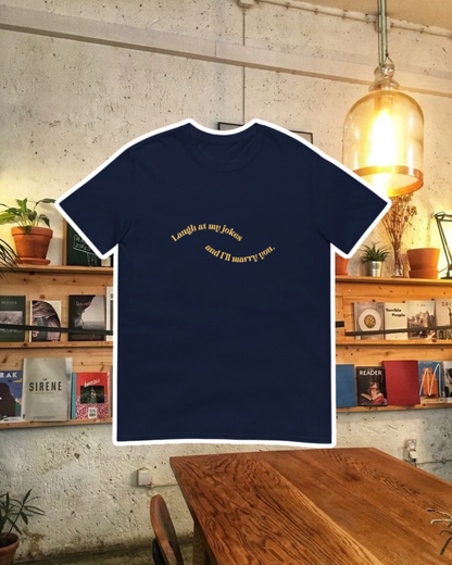 Navy T-Shirt with the phrase 'Laugh at my jokes and I will marry you' on the chest, displayed against a vibrant café-style backdrop with wood chairs and colorful cozy decoration. A playful and bold design for fans of funny merchandise and sarcasm.