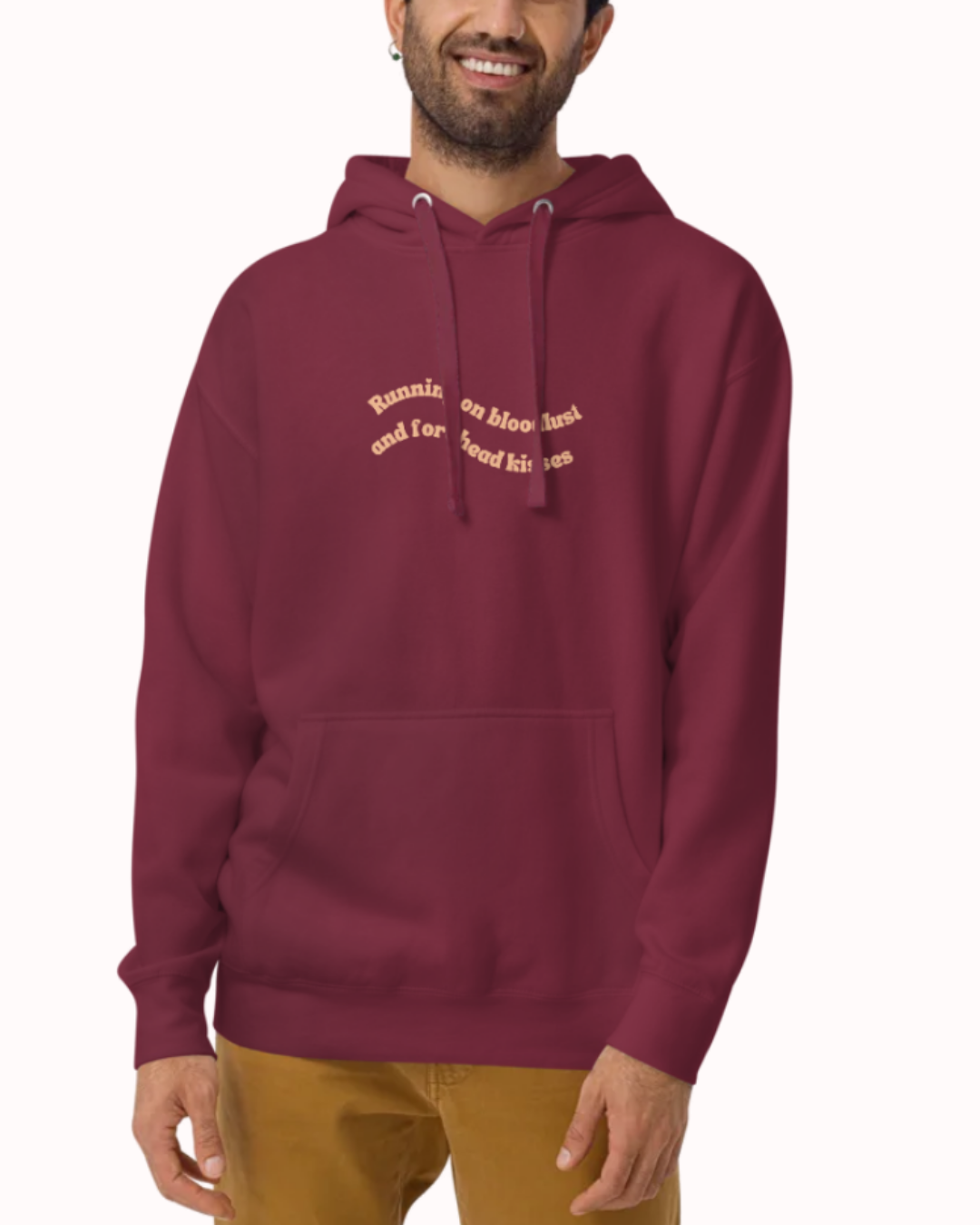 Front view of a Maroon hoodie worn by a smiling man, featuring the phrase "Running on bloodlust and forehead kisses" on the chest and back. Perfect for fans of funny merchandise, bold humor, and witty sarcasm.