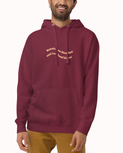 Front view of a Maroon hoodie worn by a smiling man, featuring the phrase "Running on bloodlust and forehead kisses" on the chest and back. Perfect for fans of funny merchandise, bold humor, and witty sarcasm.